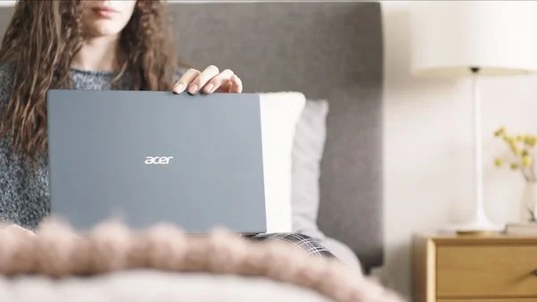 Person sitting on bed with gray Acer laptop open on lap, in a bedroom with a nightstand, lamp, and plant in the background.