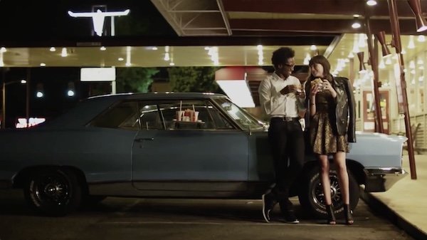 A vintage blue car parked in front of a lit-up building at night, with a man and woman standing next to it looking at their phones.