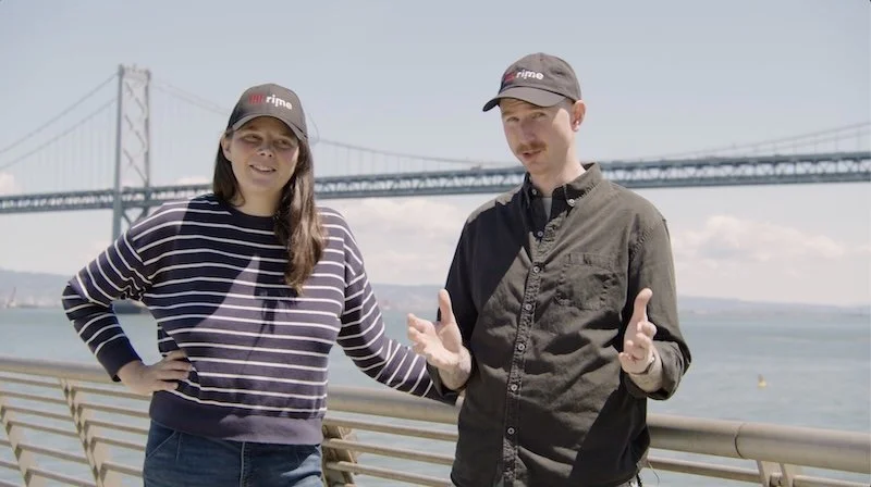 Two people standing near a river with a large suspension bridge in the background, wearing caps and casual clothing, engaging in conversation.