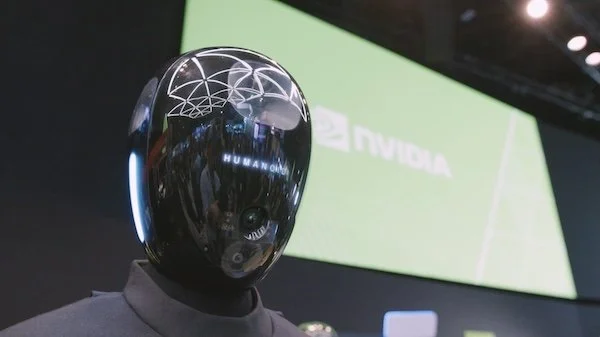 A humanoid robot with a shiny black head, displaying the word 'HUMANOID' on its face, at a technology event with a green 'NVIDIA' screen in the background.