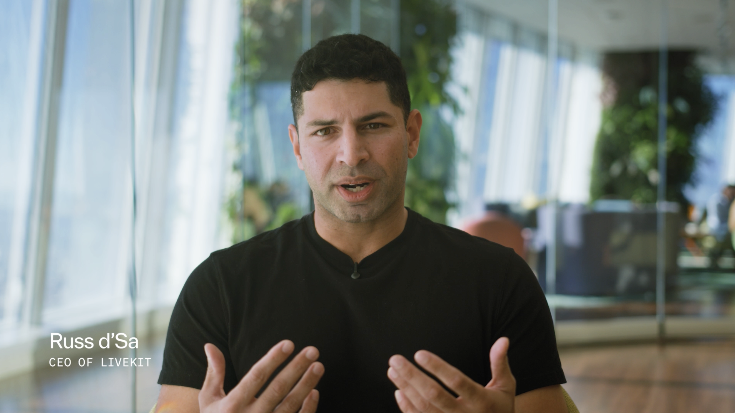 Man in a suit sitting in a modern office with large windows and plants, San Francisco skyline visible.