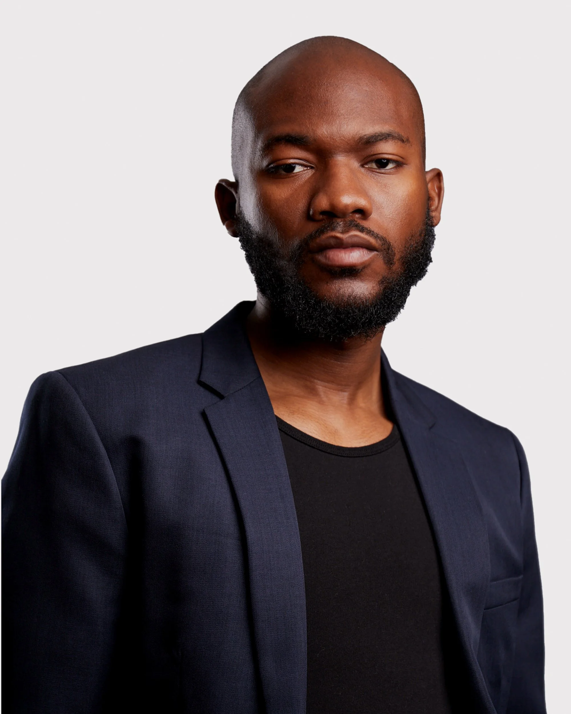 Close-up of a man with a shaved head and a beard, wearing a dark blazer over a black shirt, against a plain light background.