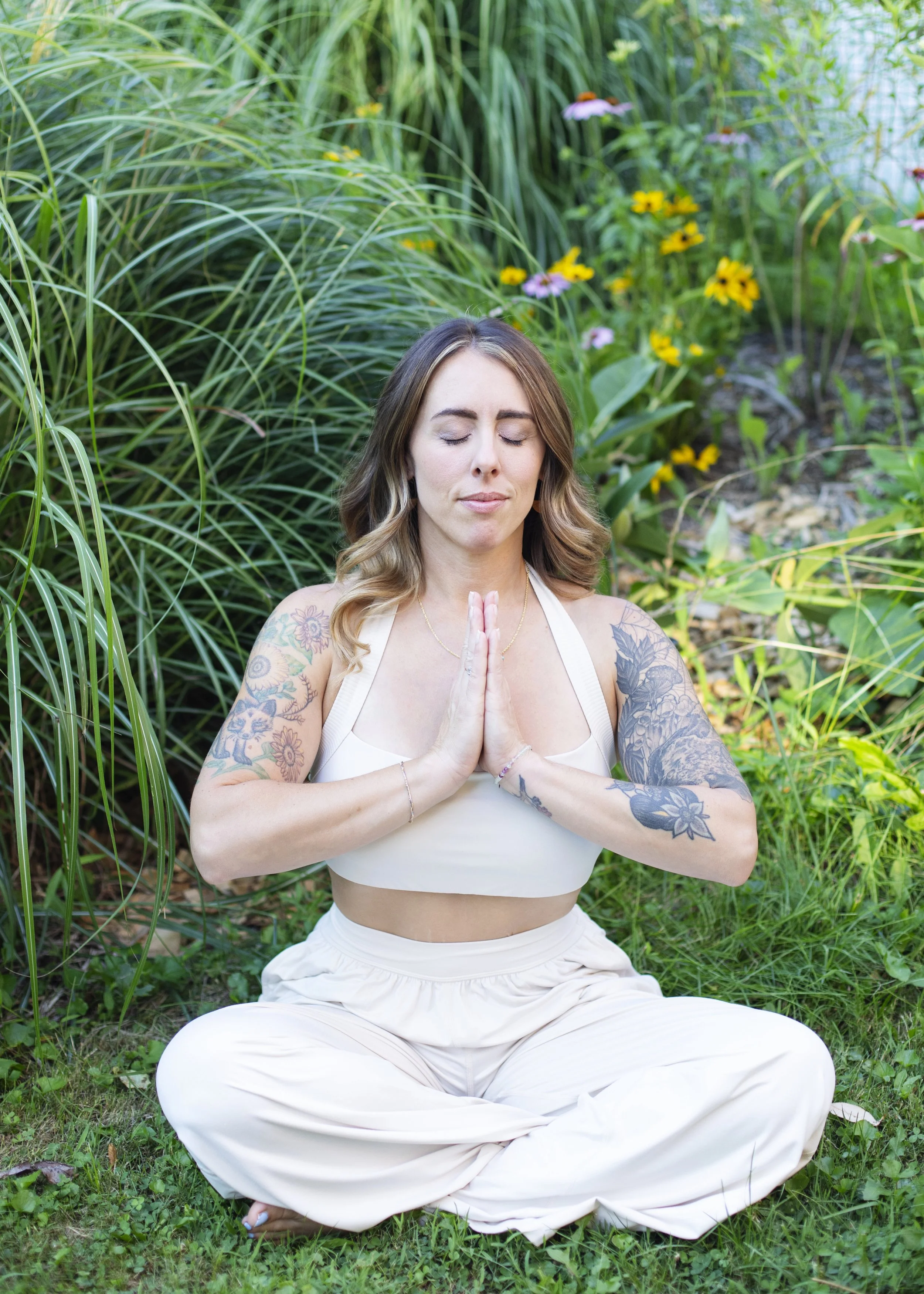 A woman practicing yoga outdoors with eyes closed, sitting cross-legged on grass, hands in prayer position, surrounded by green plants and colorful flowers.