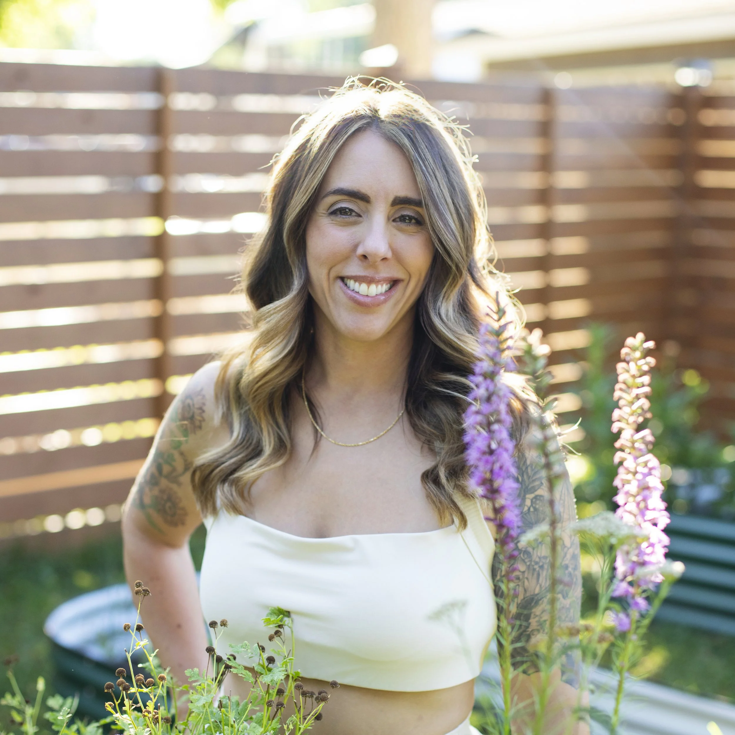 A smiling woman with long wavy hair and tattoos on her arm, standing outdoors in a garden with a wooden fence in the background.