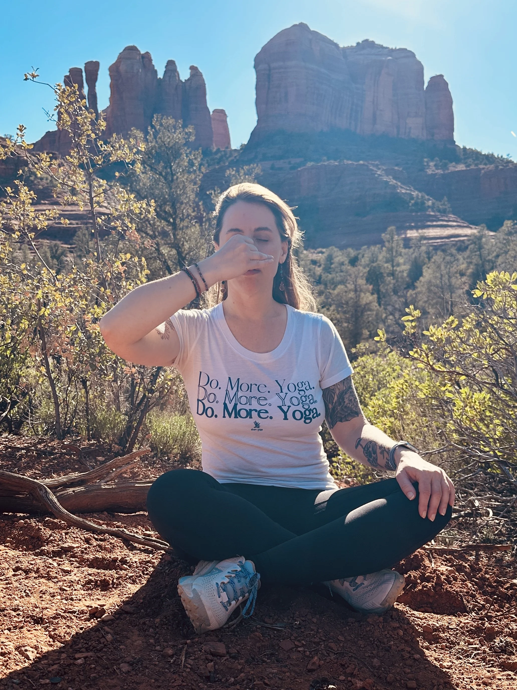 A woman sitting cross-legged on the ground outdoors with a mountain background, wearing a white T-shirt with the text 'Do. More. Yoga.' and black pants, holding her nose with her right hand in a yoga pose.