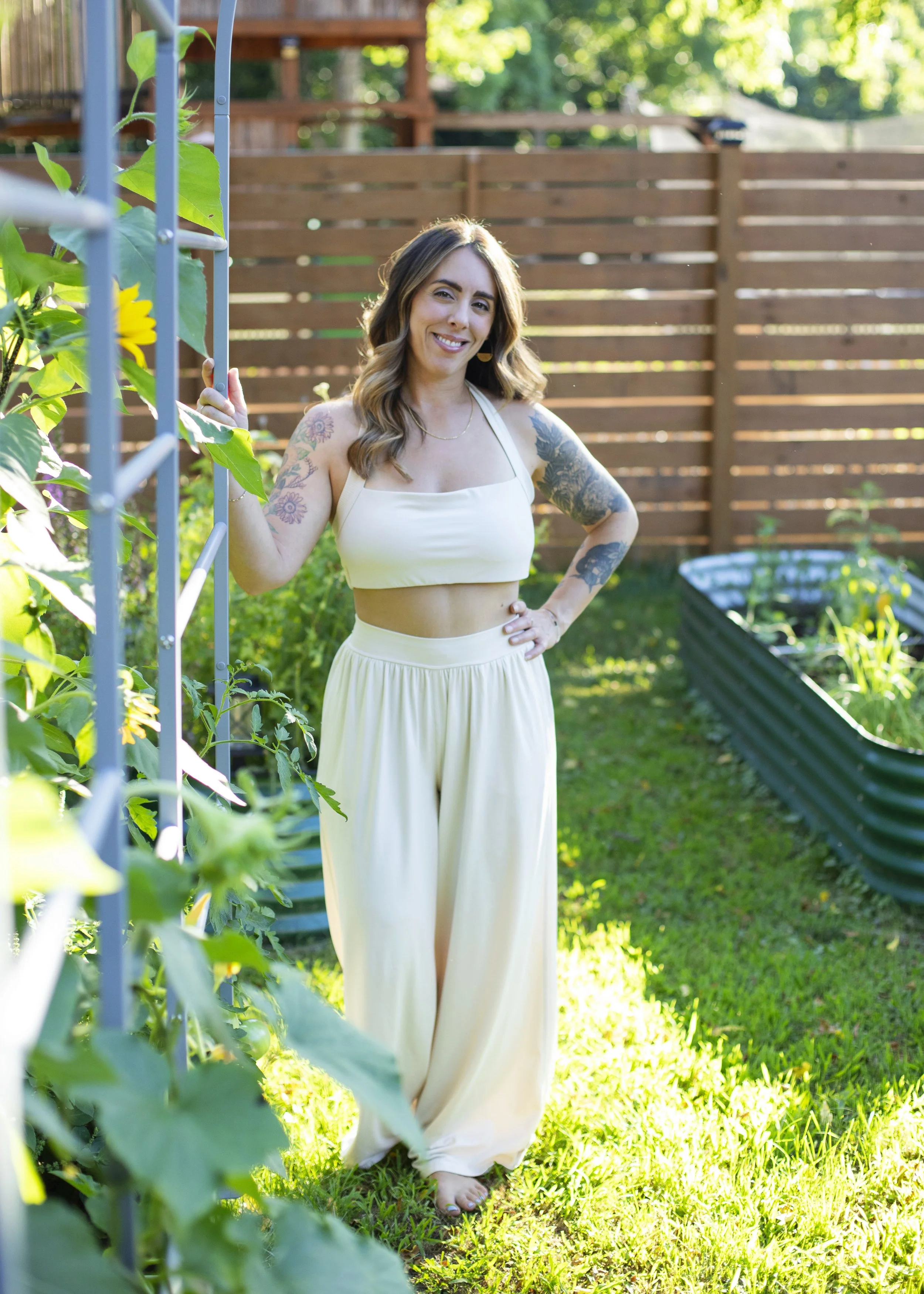 A woman with tattoos on her arms standing barefoot in a garden, smiling, wearing a white crop top and cream-colored wide-leg pants, holding a metal garden frame with green leaves and yellow flowers, with a wooden fence and greenery in the background.