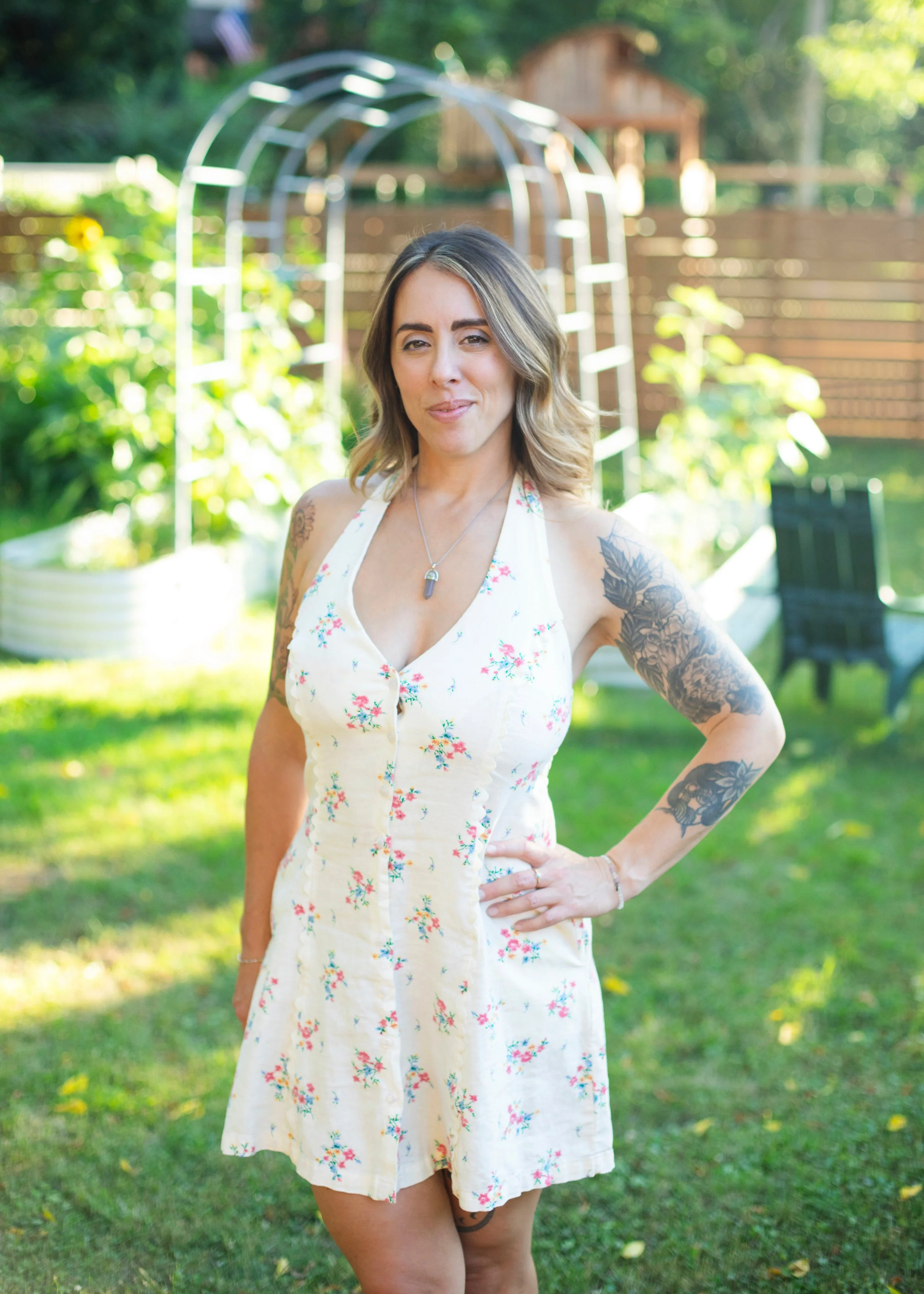A woman with tattoos standing outdoors in a garden with sunlight, wearing a light-colored floral dress, smiling and winking at the camera.