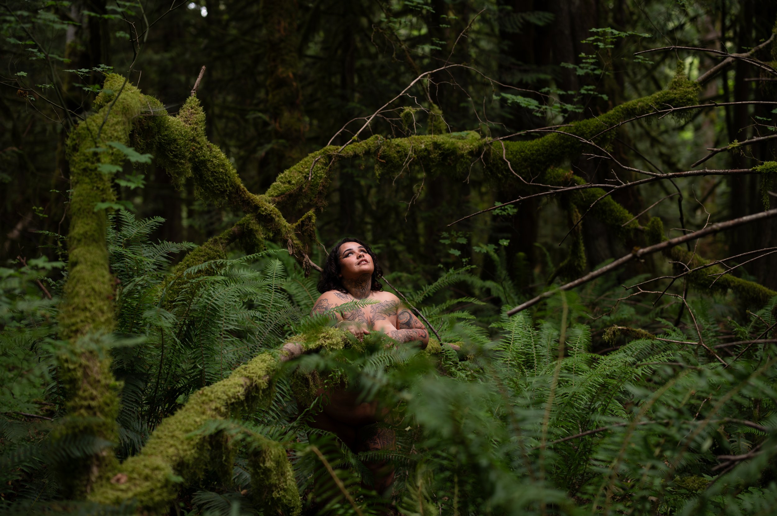 Nude person rests her arms on a mossy tree branch and is surrounded by ferns
