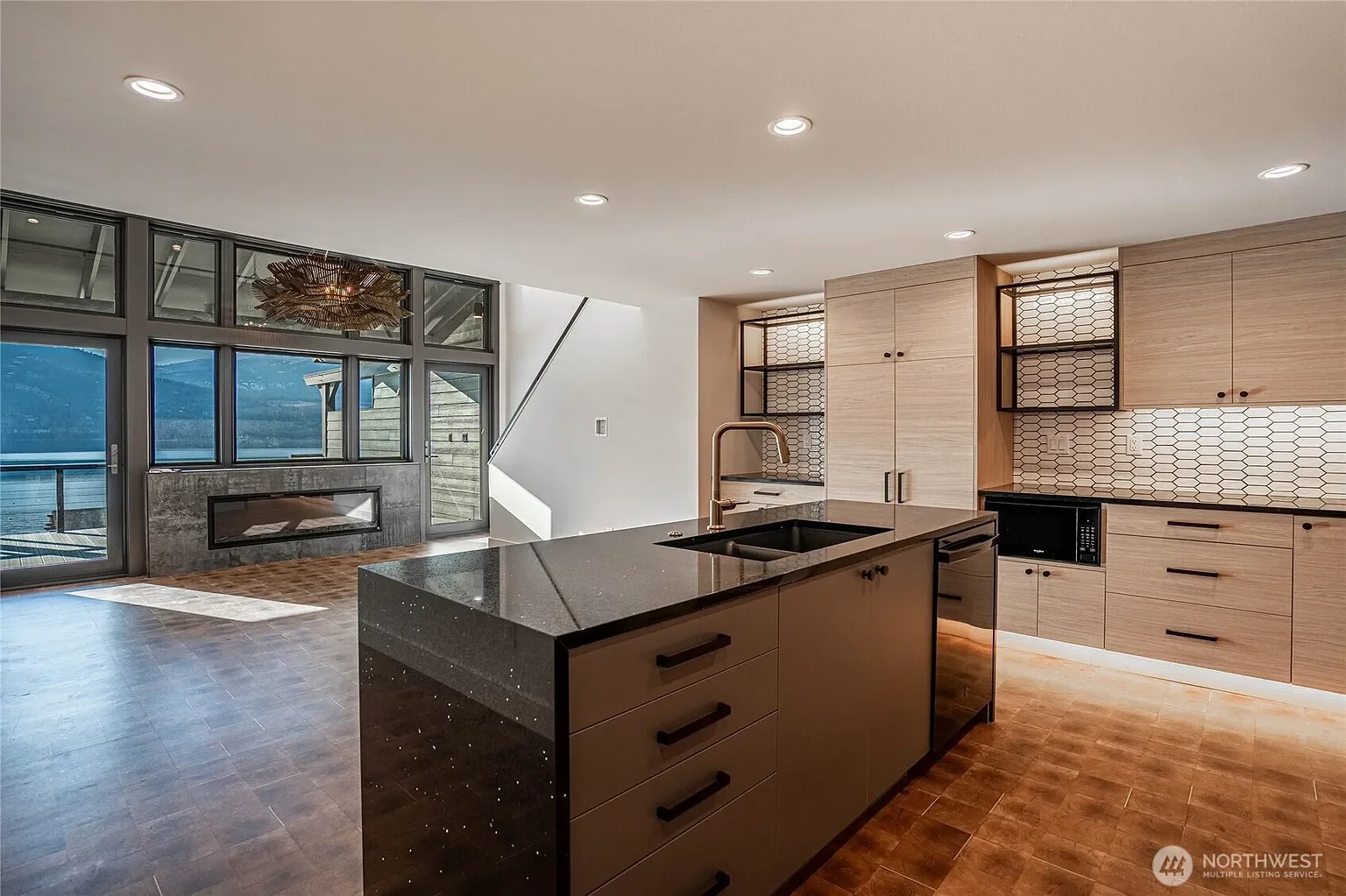Modern kitchen with black marble countertop, light wood cabinets, black appliances, hexagonal tile backsplash, and a large island with a wooden bar.