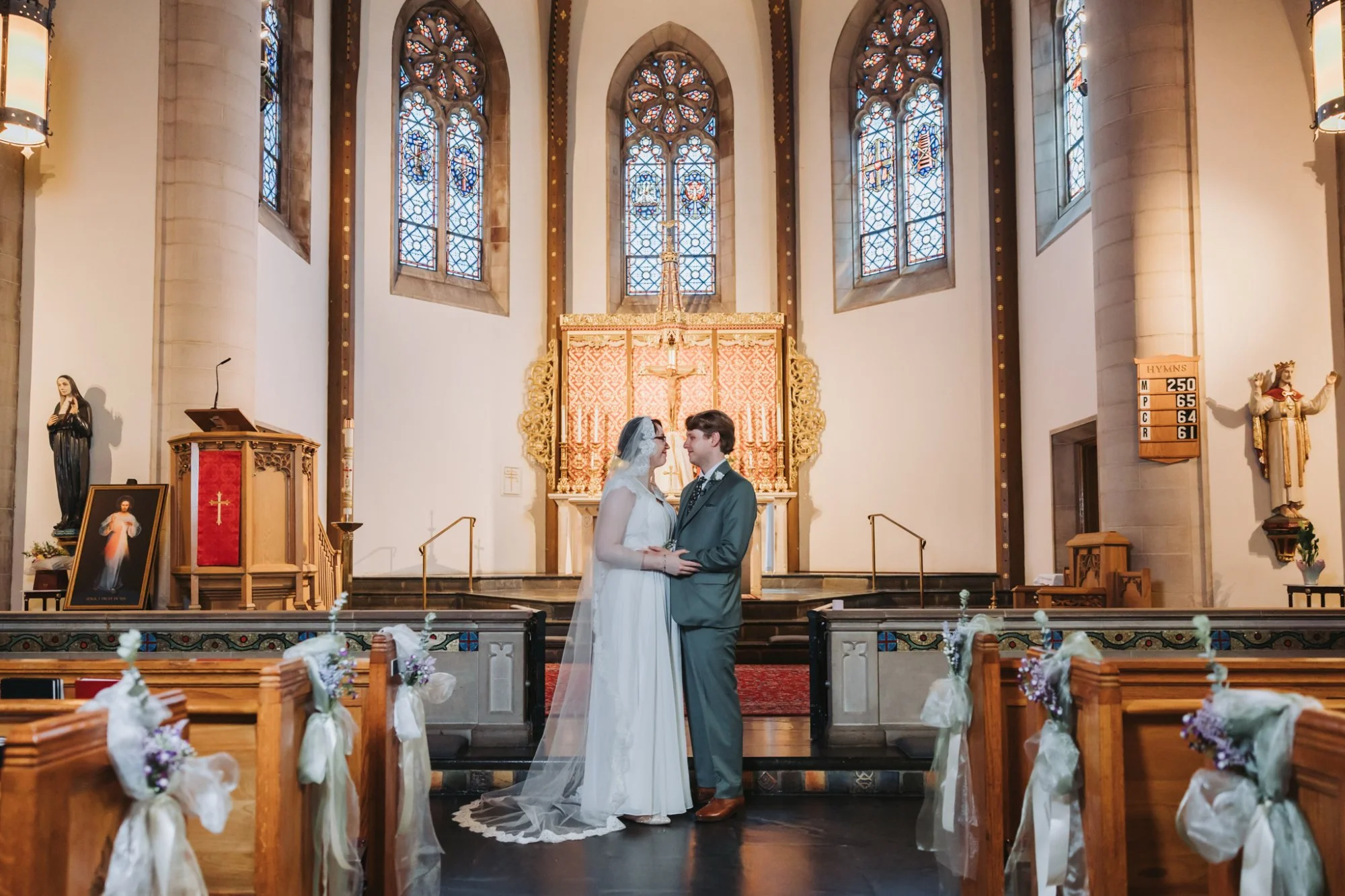 A bride and groom standing face-to-face inside a church, holding hands. The church has stained glass windows and an ornate altar behind them. The pews are decorated with flowers and ribbons.