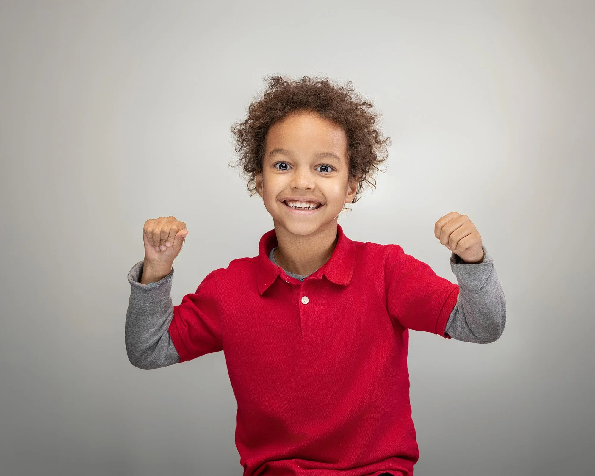 A young boy with curly hair smiling and showing his teeth, wearing a red polo shirt and gray long-sleeve shirt underneath, raising his arms with fists clenched in a gesture of strength.