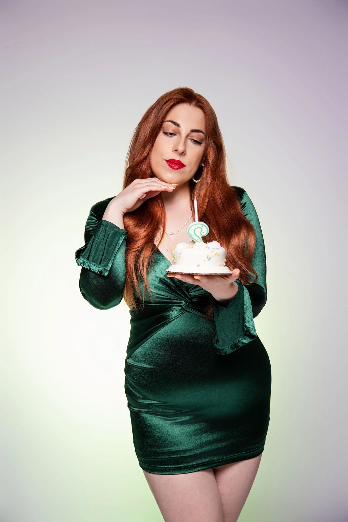 A woman with long red hair wearing a dark green, shiny dress holding a small birthday cake with a lit candle, looking at it thoughtfully.
