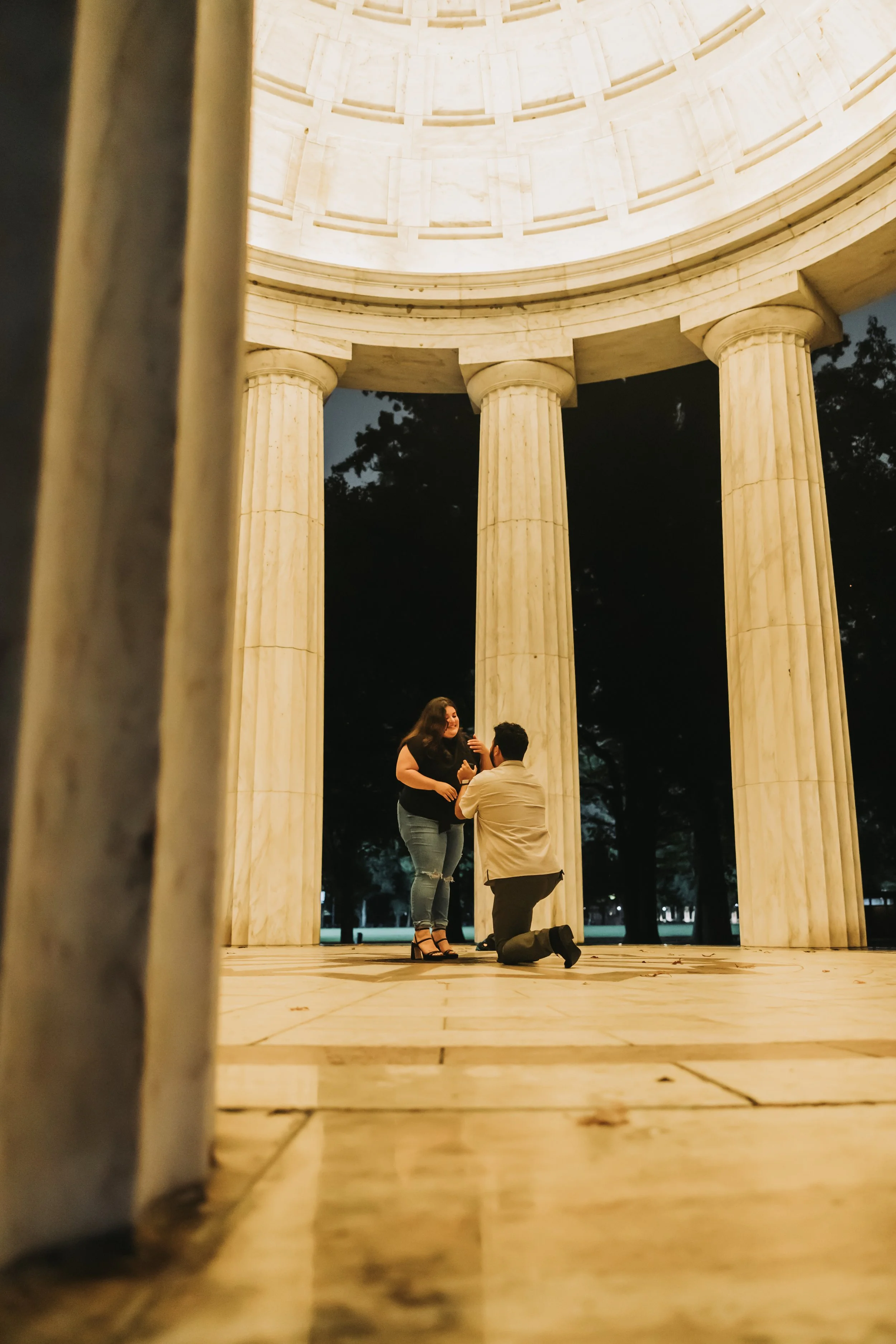 A man proposing to a woman at night inside the DC War Memorial, seen from a low angle.