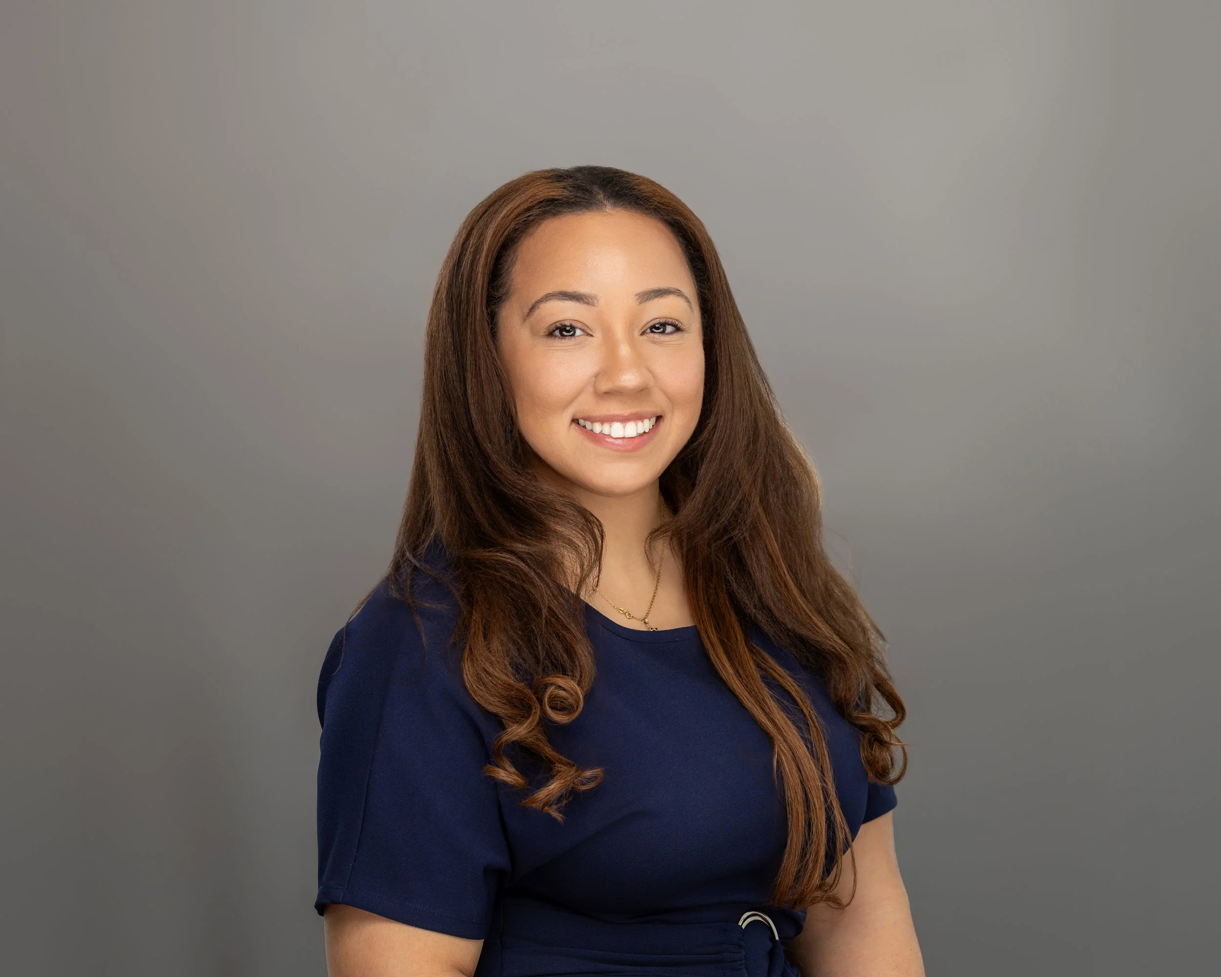 A young woman with long, wavy brown hair, smiling, wearing a navy blue dress, standing against a plain gray background.
