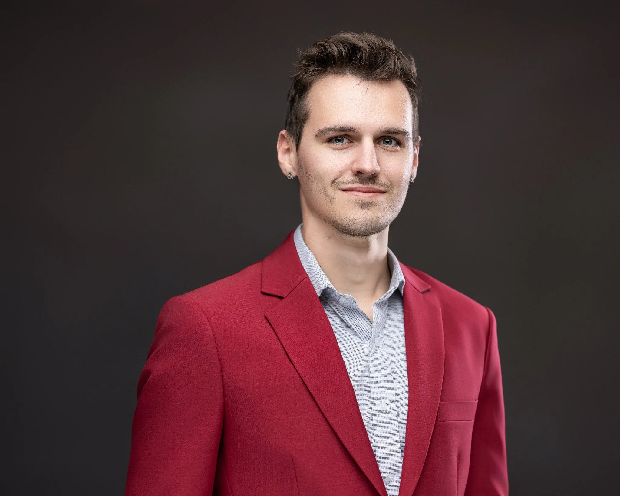 Portrait of a young man with fair skin, wearing a red blazer and a light gray collared shirt, against a dark background.