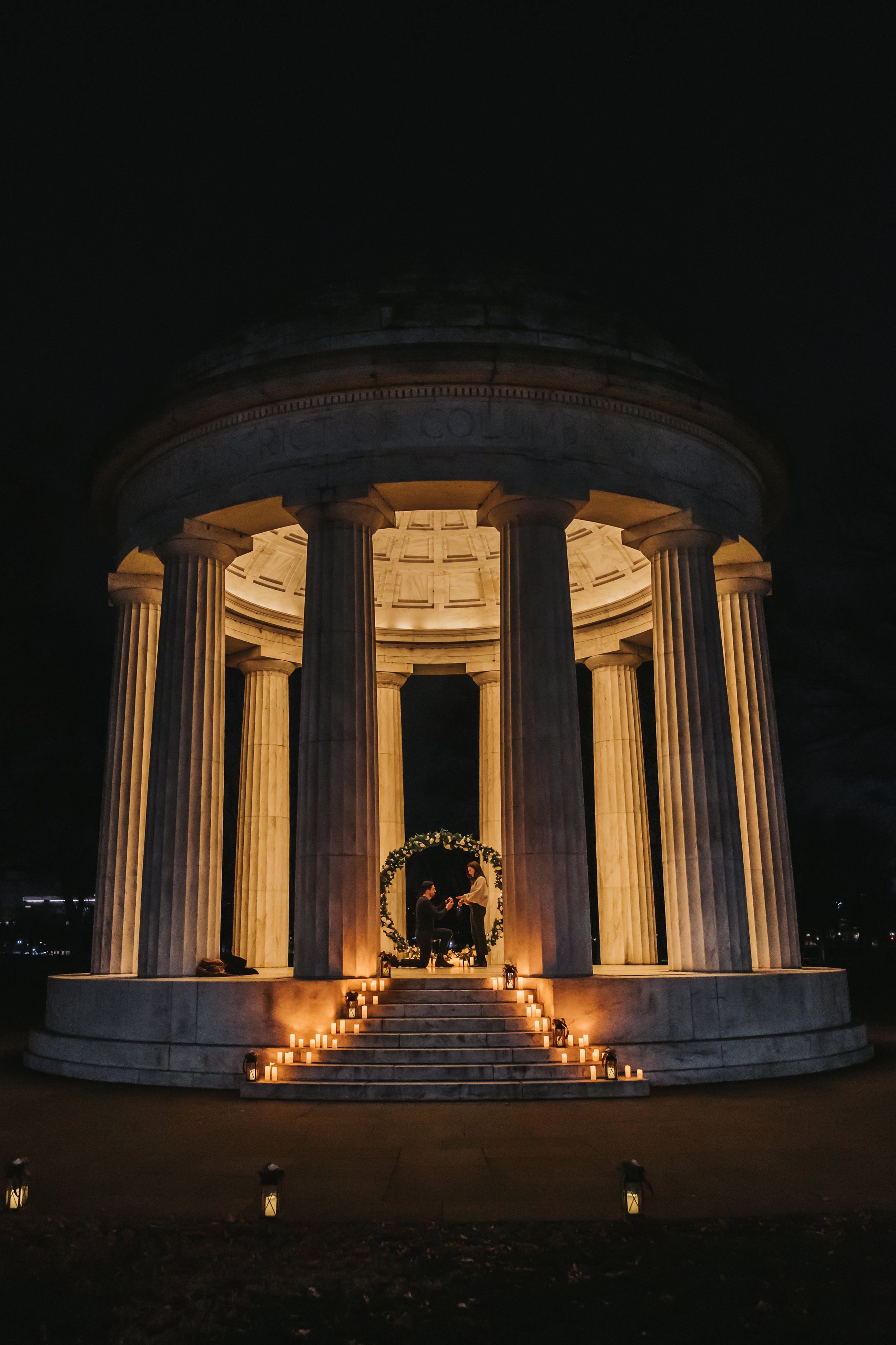 A nighttime scene at the DC War Memorial with an illuminated circular structure featuring tall columns. In front of the structure, a man proposes to a woman surrounded by candles on the steps.