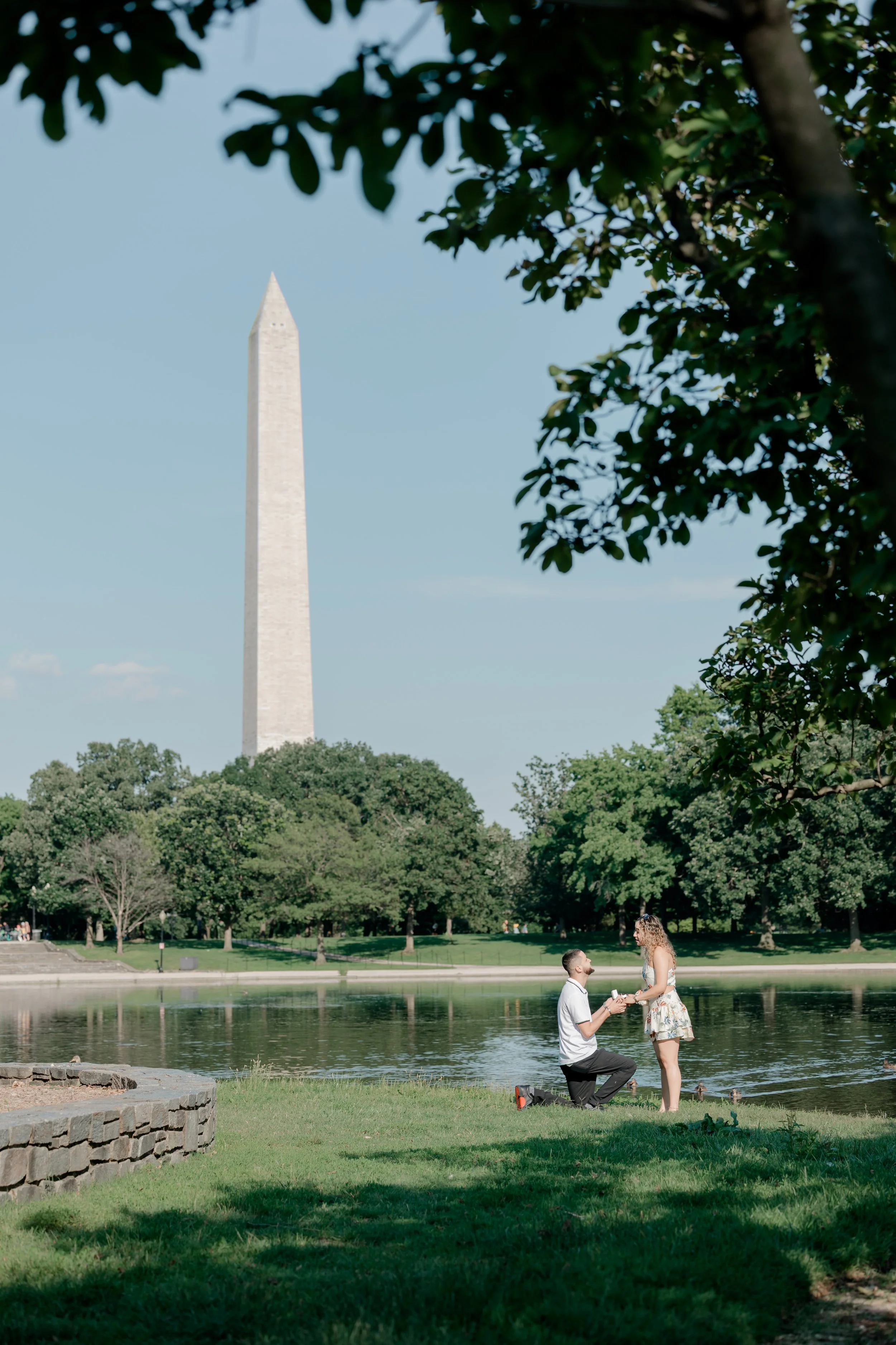 Man proposing to girlfriend in grass at Constitution Gardens with a view of the Washington Monument.
