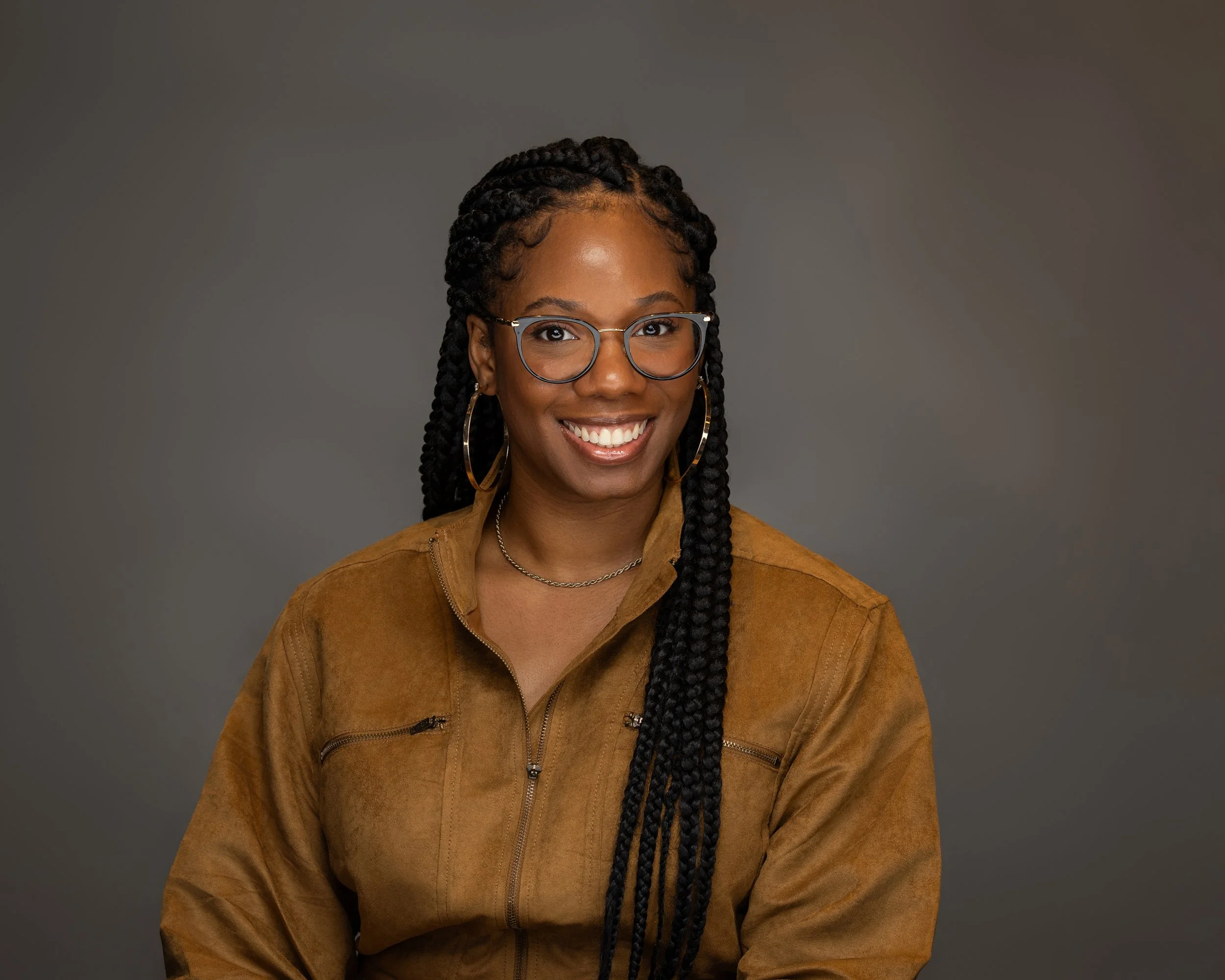 Portrait of a woman with glasses, large hoop earrings, and long braided hair, wearing a brown suede jacket, smiling against a gray background.
