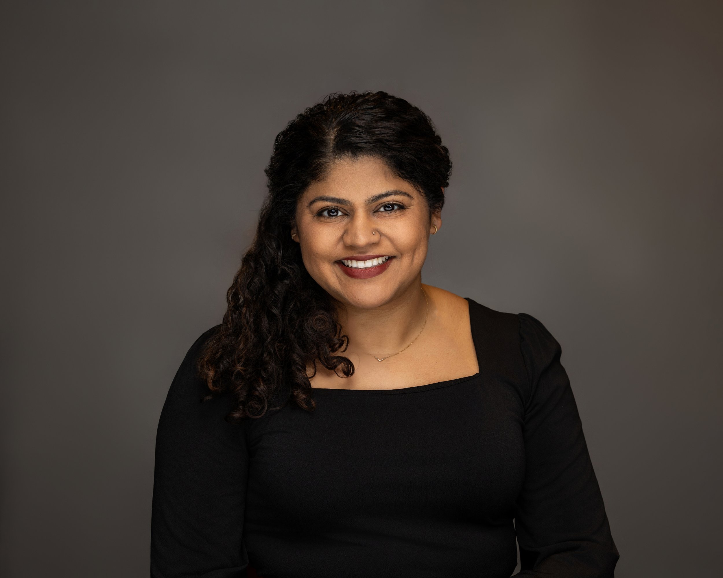 Portrait of a smiling woman with dark curly hair wearing a black top against a gray background.