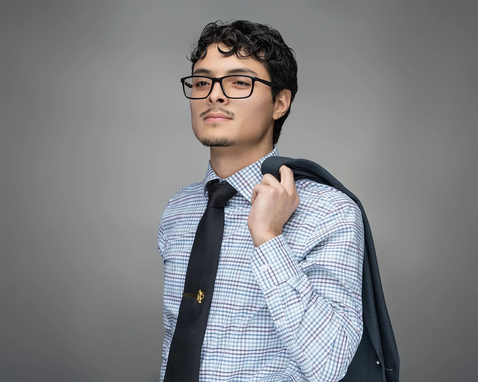 A young man with glasses wearing a checkered shirt and black tie, holding a jacket over his shoulder, standing against a plain gray background.