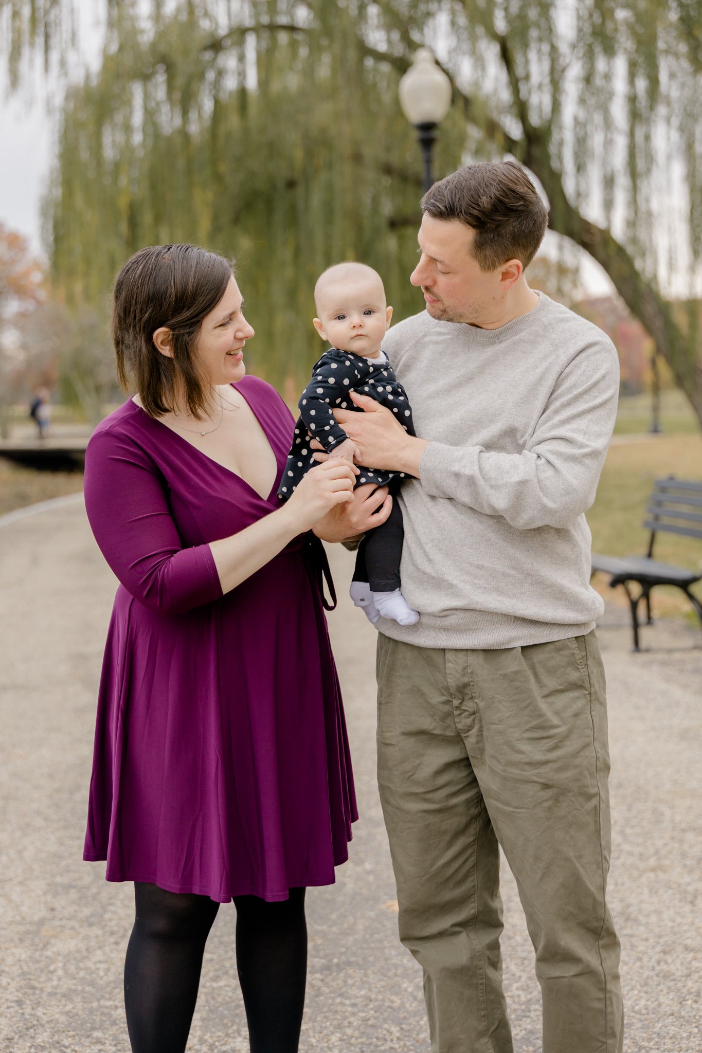 Family of two adults and a baby sitting on a park bench with their dog, surrounded by autumn trees with orange and green leaves.