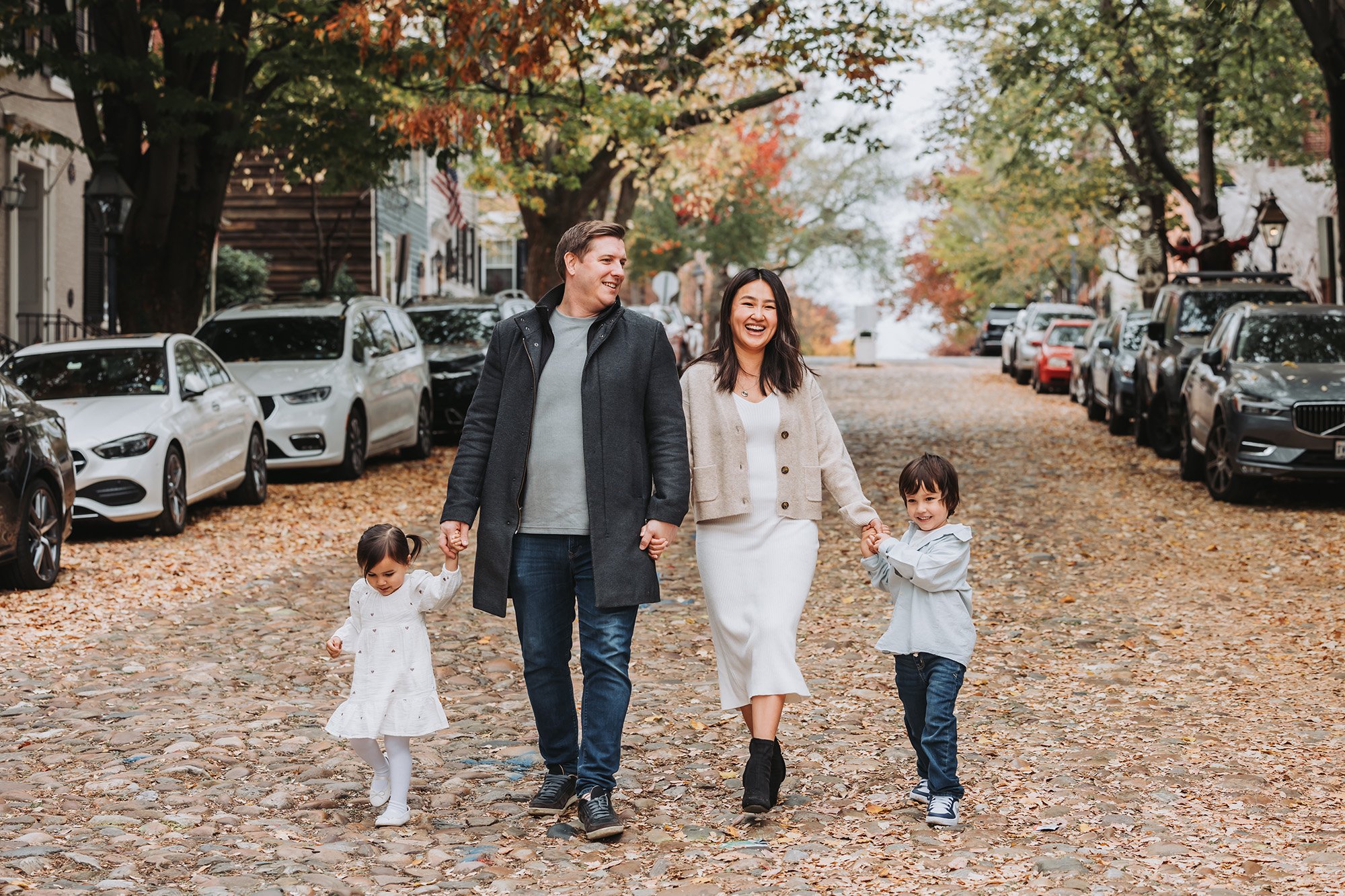 A family of four walking hand in hand on a cobblestone street in autumn, surrounded by parked cars and trees with colorful fall leaves.