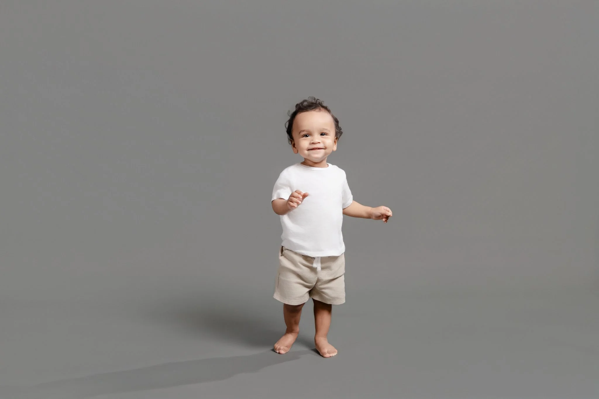A young child with short curly dark hair, wearing a white t-shirt and beige shorts, standing barefoot on a plain gray background, smiling and looking at the camera.