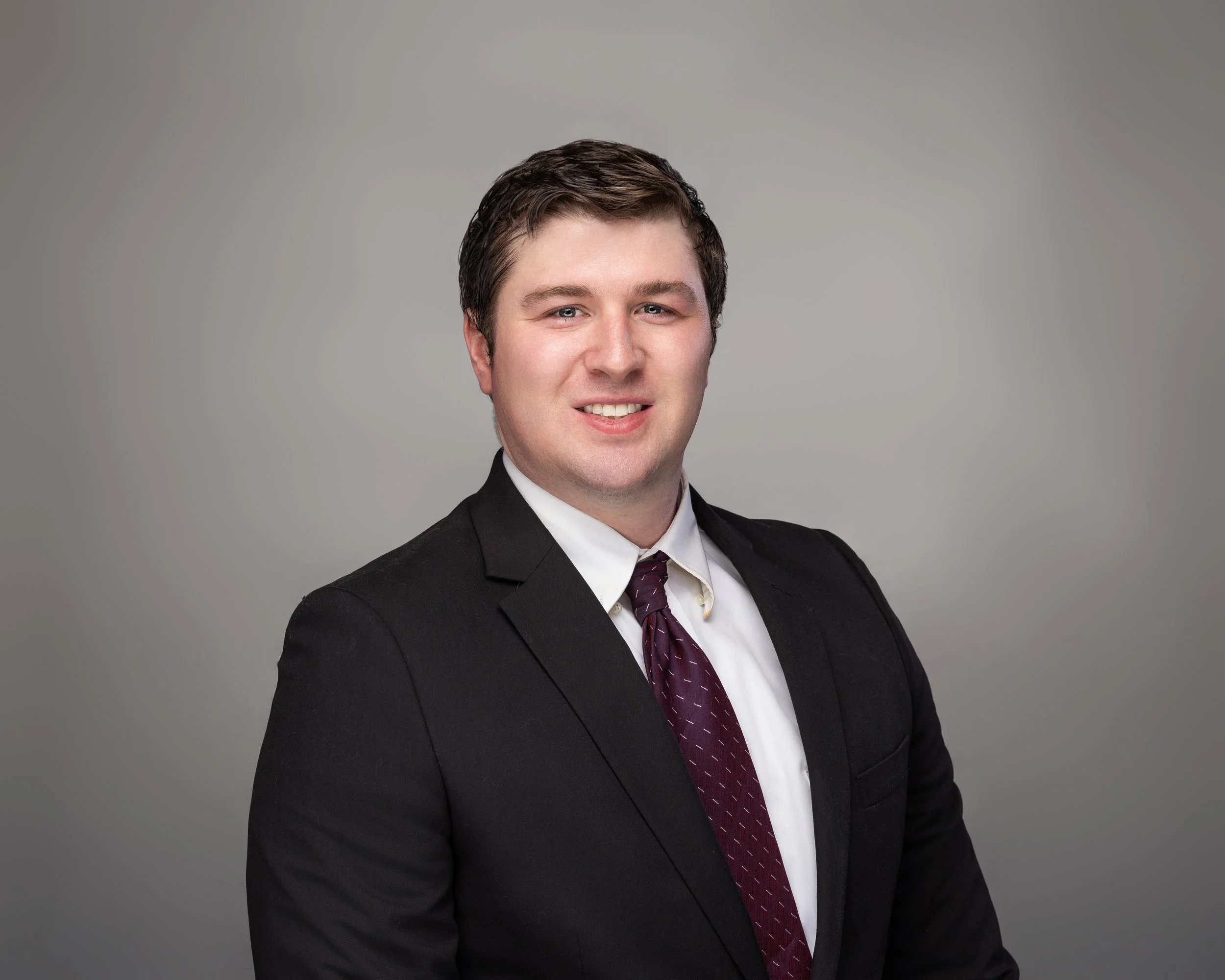 Portrait of a young man with dark hair wearing a black suit, white shirt, and maroon tie, smiling against a plain gray background.