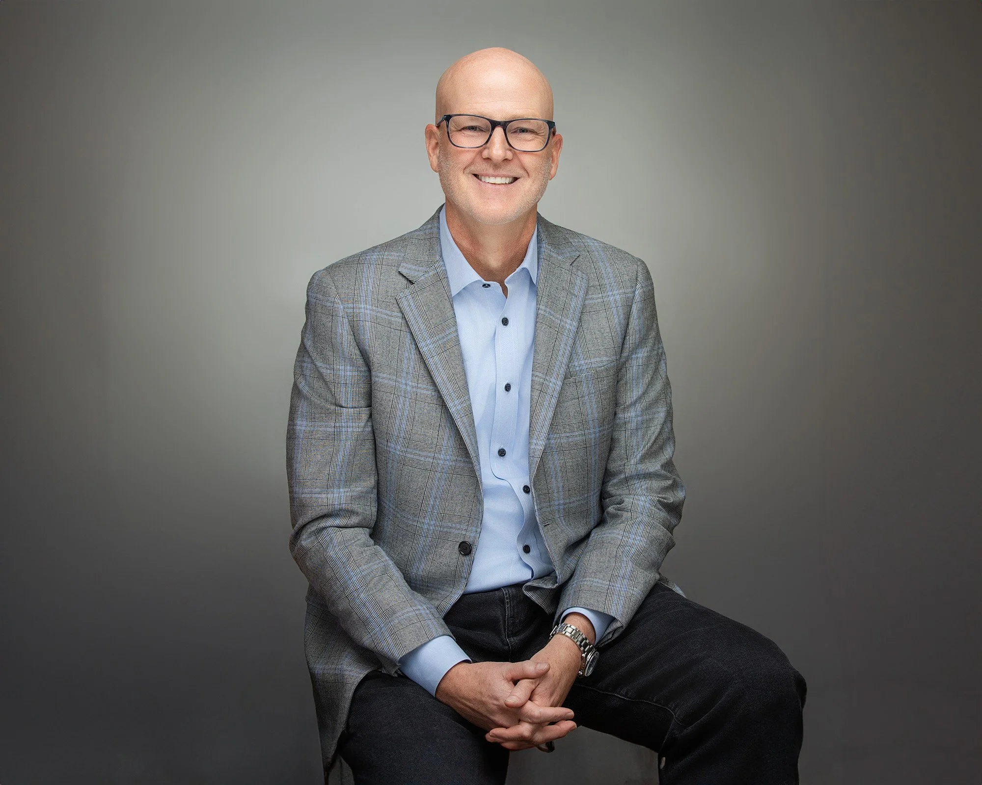 A middle-aged man with glasses, wearing a gray plaid blazer with a light blue shirt, sitting against a plain gray background, smiling at the camera.