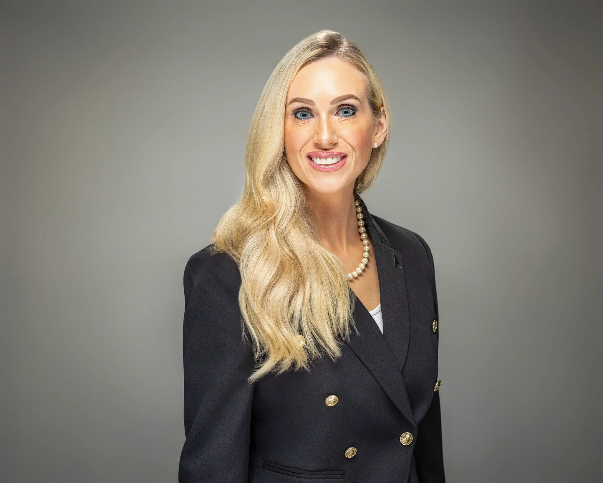 A woman with long blonde hair, wearing a black blazer with gold buttons, a pearl necklace, and a white top, smiling against a gray background.