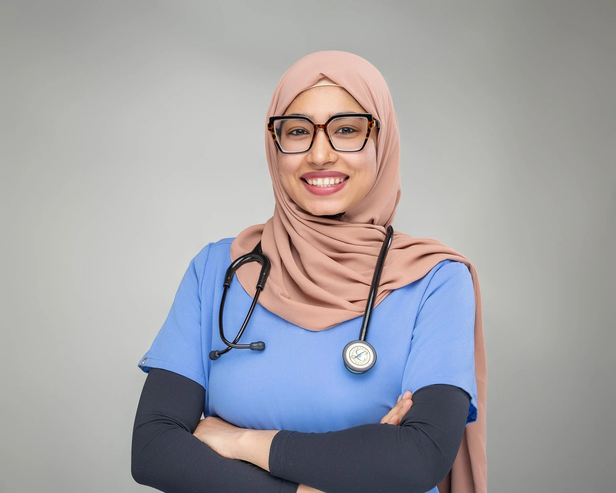 A smiling female healthcare professional wearing glasses and a hijab, with a stethoscope around her neck, crossed arms, and dressed in a blue medical uniform against a plain gray background.