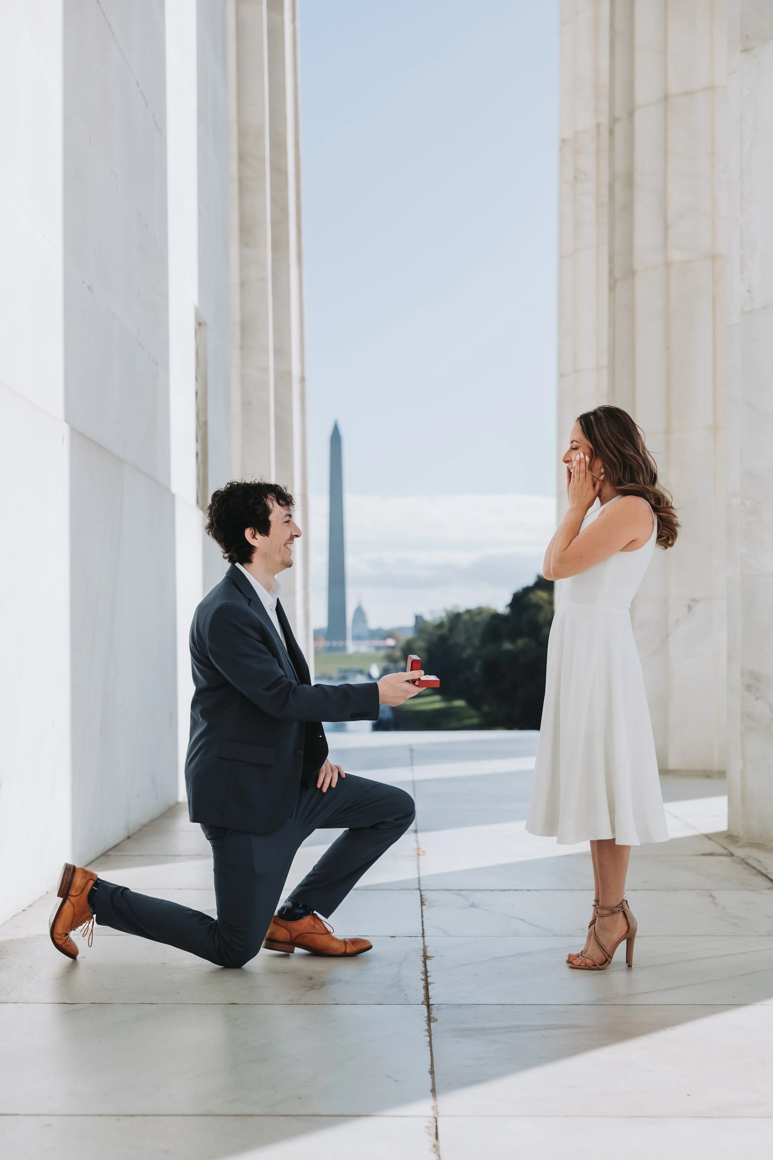 A man on one knee holding a ring box, proposing to a woman in a white dress at the Lincoln Memorial, the Washington Monument visible in the background.