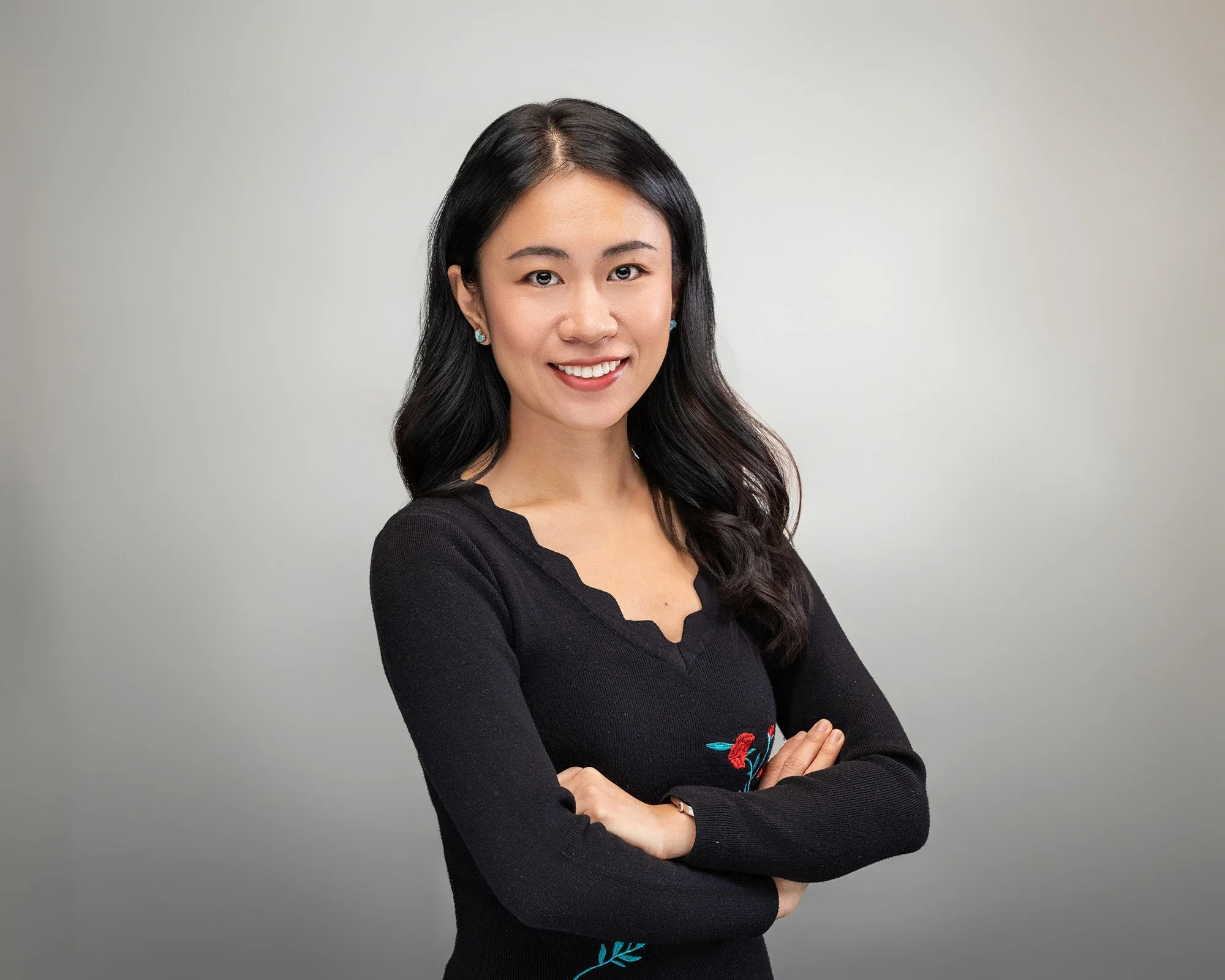 A woman with black hair, smiling and crossing her arms, wearing a black top with embroidery, standing against a plain gray background.