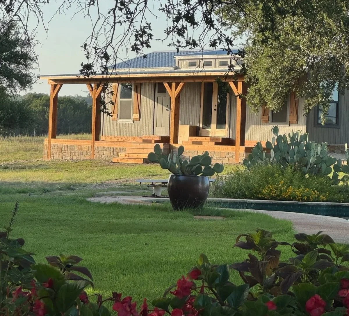 A house with a wooden porch, steps, and a metal roof, surrounded by greenery and cacti in a large pot, with a landscaped yard and flowering plants.