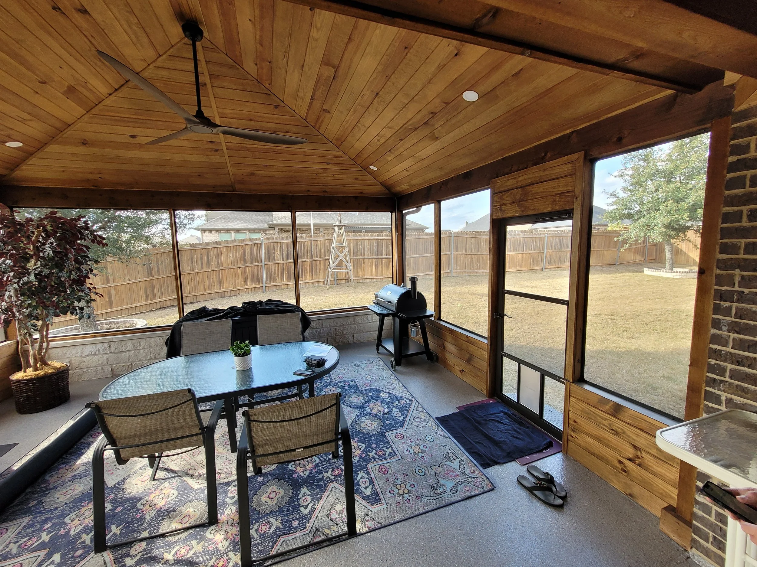 Enclosed patio with wooden ceiling, ceiling fan, table with four chairs, potted plant, small black grill, and slippers on the floor, overlooking a backyard with a fence, trees, and a ladder.