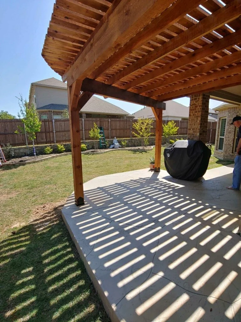 Backyard patio area with a wooden pergola casting striped shadows, a covered barbecue grill, and a person standing nearby. There are small trees and plants along a fenced yard with neighboring houses in the background.