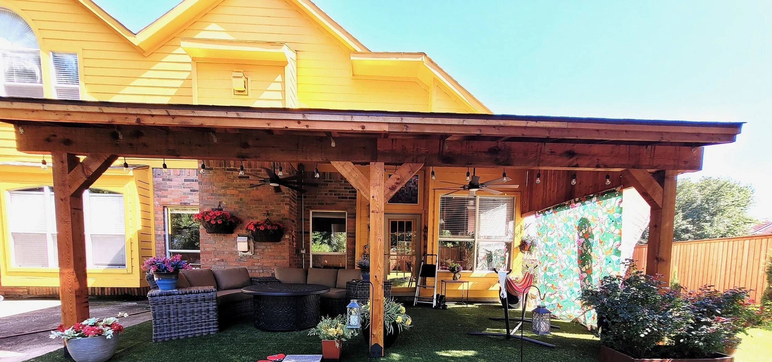 Backyard patio with wooden pergola, outdoor furniture, potted plants, hanging flower baskets, and a decorative fabric wall panel. Yellow house with brick accent wall in the background.