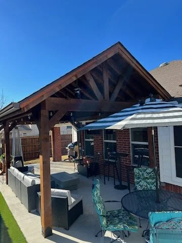 Outdoor patio with a wooden pergola, cushioned seating, metal chairs with patterned cushions, patio table, and bar-style seating along a brick wall.
