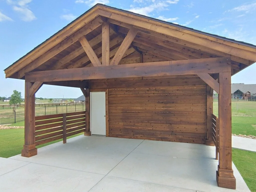 A wooden outdoor structure with a sloped roof, a side door, and a surrounding wood railing on a concrete patio, overlooking a grassy yard and outdoor landscape.