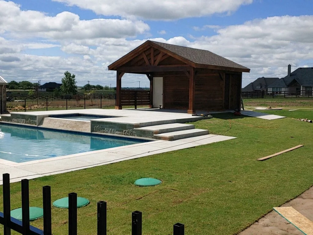A backyard with a swimming pool, a wooden pool house or shed, and a lush green lawn under partly cloudy skies.