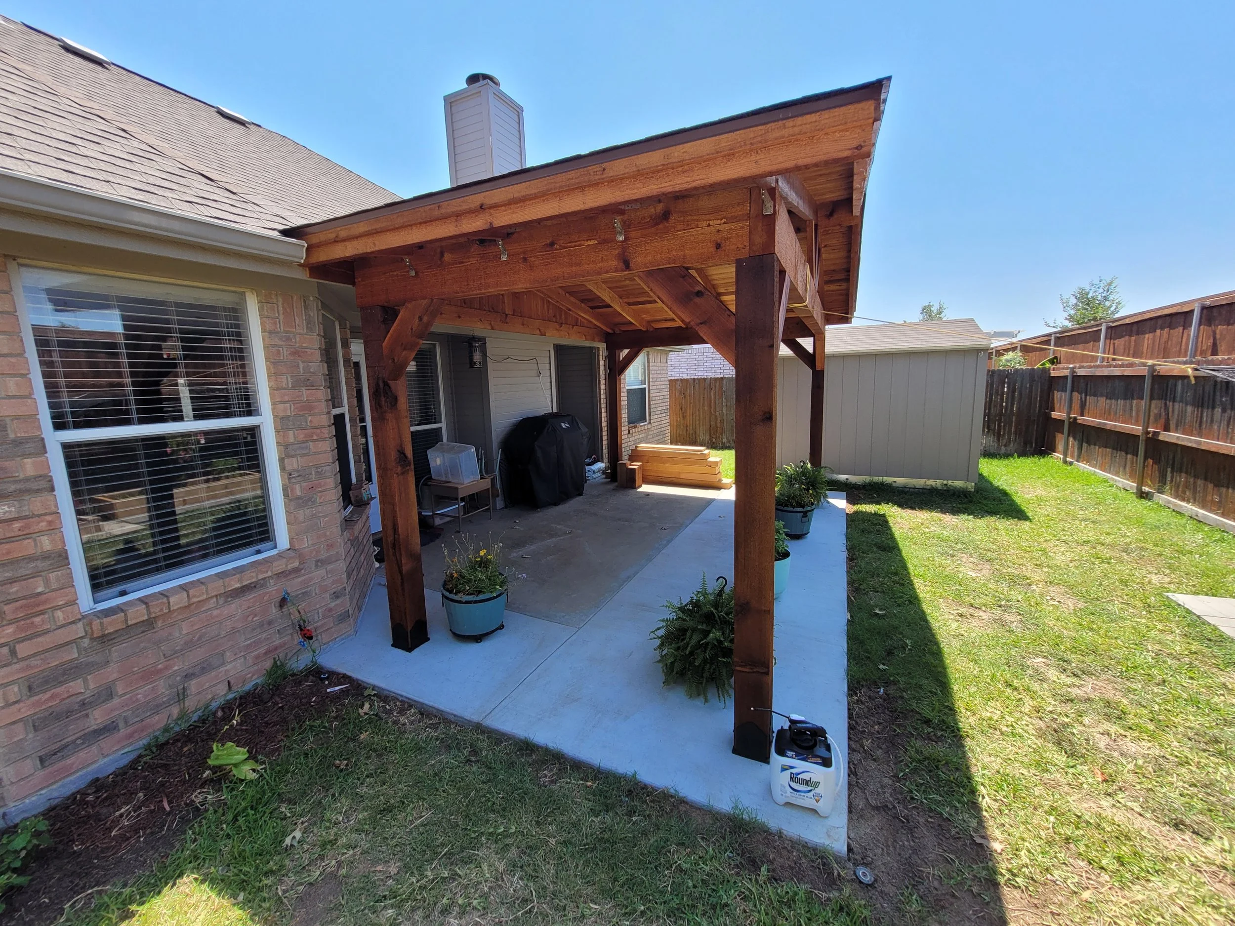 Backyard patio with a newly built wooden pergola, potted plants, a barbecue grill, and a small shed. The patio is partially shaded, and there is a bright blue sky.