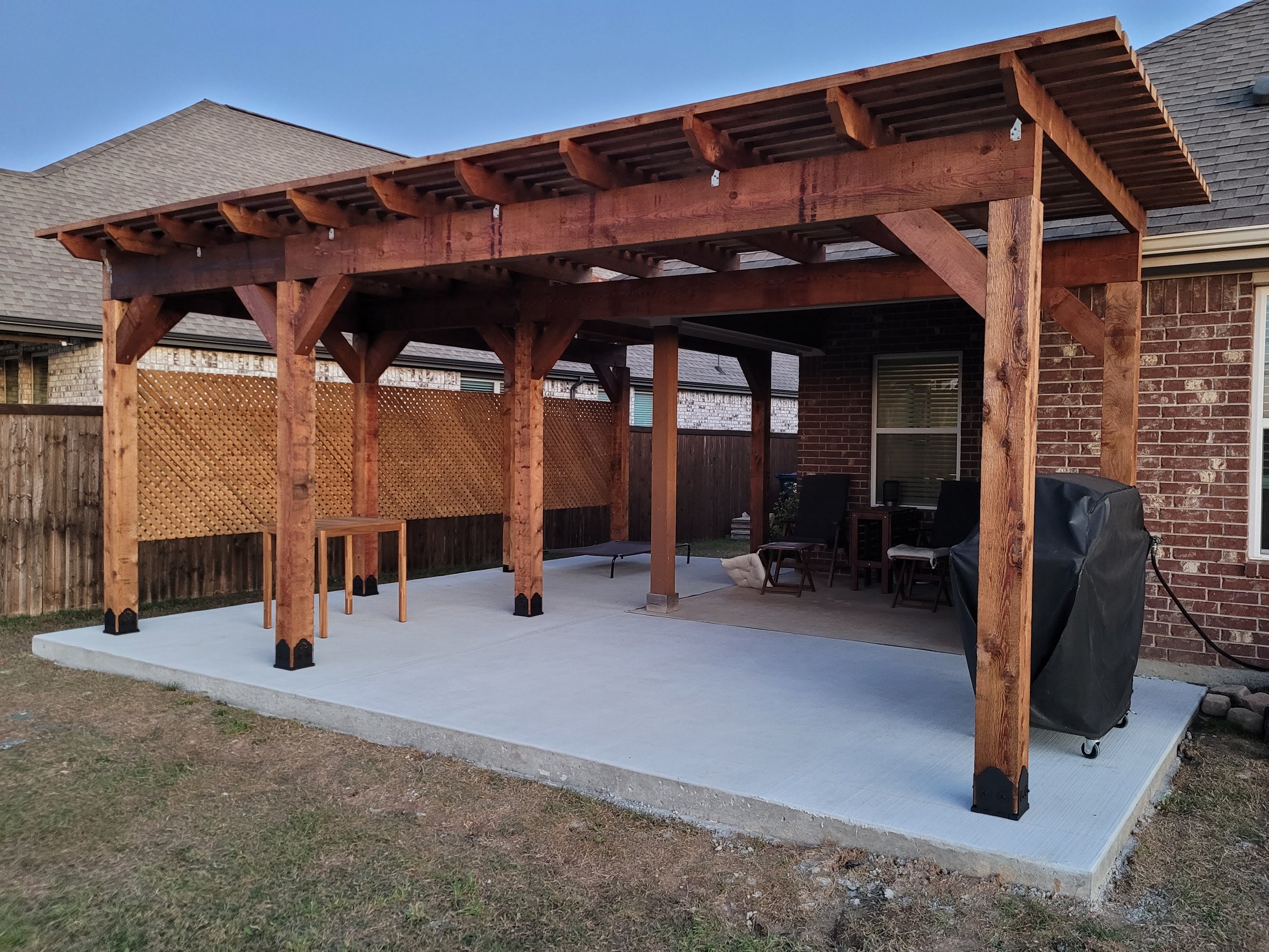 Wooden pergola structure over concrete patio next to brick house with chairs, small table, and covered grill.