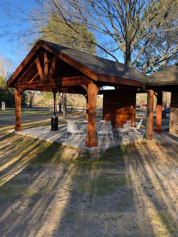 Wooden pavilion with outdoor tables and chairs beneath, casting long shadows on the ground, with leafless trees and a clear blue sky in the background.
