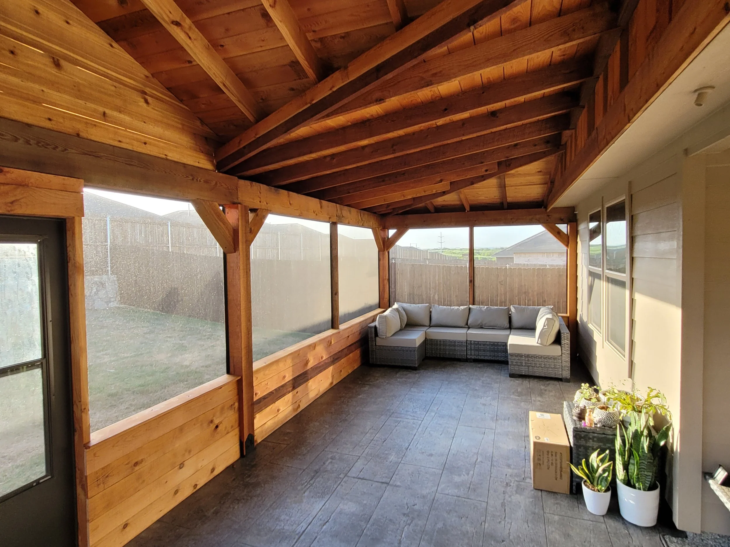 A covered outdoor patio with wooden beams and ceiling, a corner sectional sofa with light cushions, potted plants, and a cardboard box, with a view of a grassy yard and residential fencing in the background.