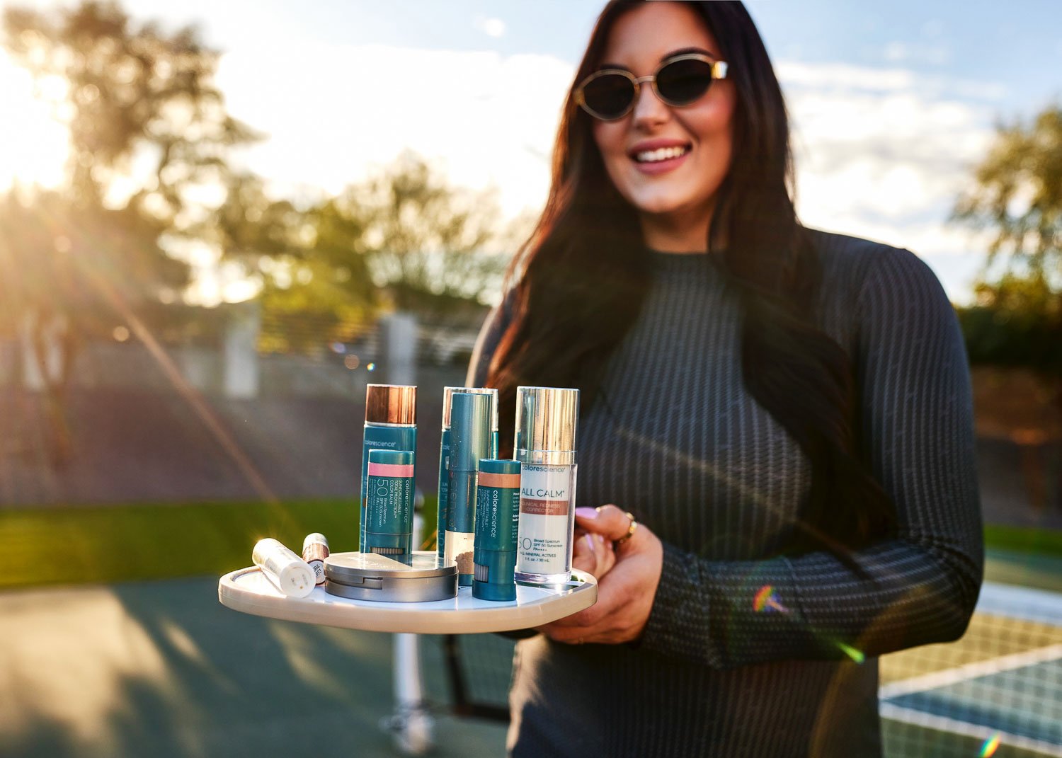 Woman holding a tray of Colorscience mineral sunscreen products outdoors at a tennis court near a VI peel provider in Peoria AZ.