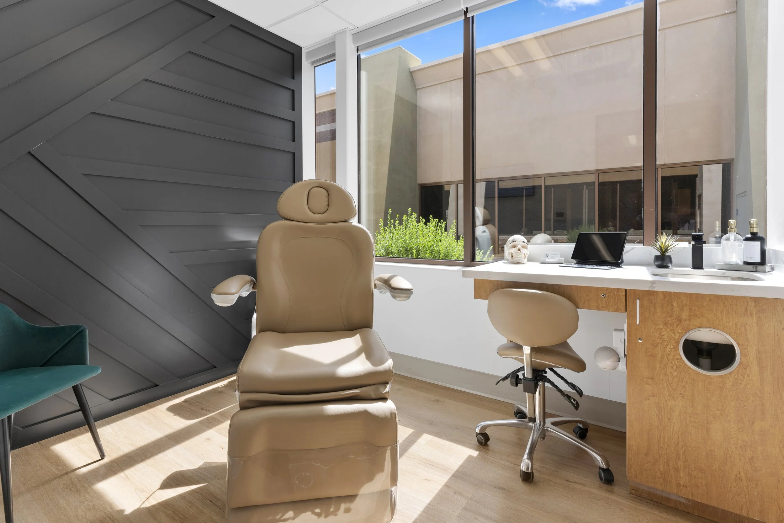 Modern office with beige leather chair, desk with laptop, skull model, plant, and decor, large windows showing buildings outside.
