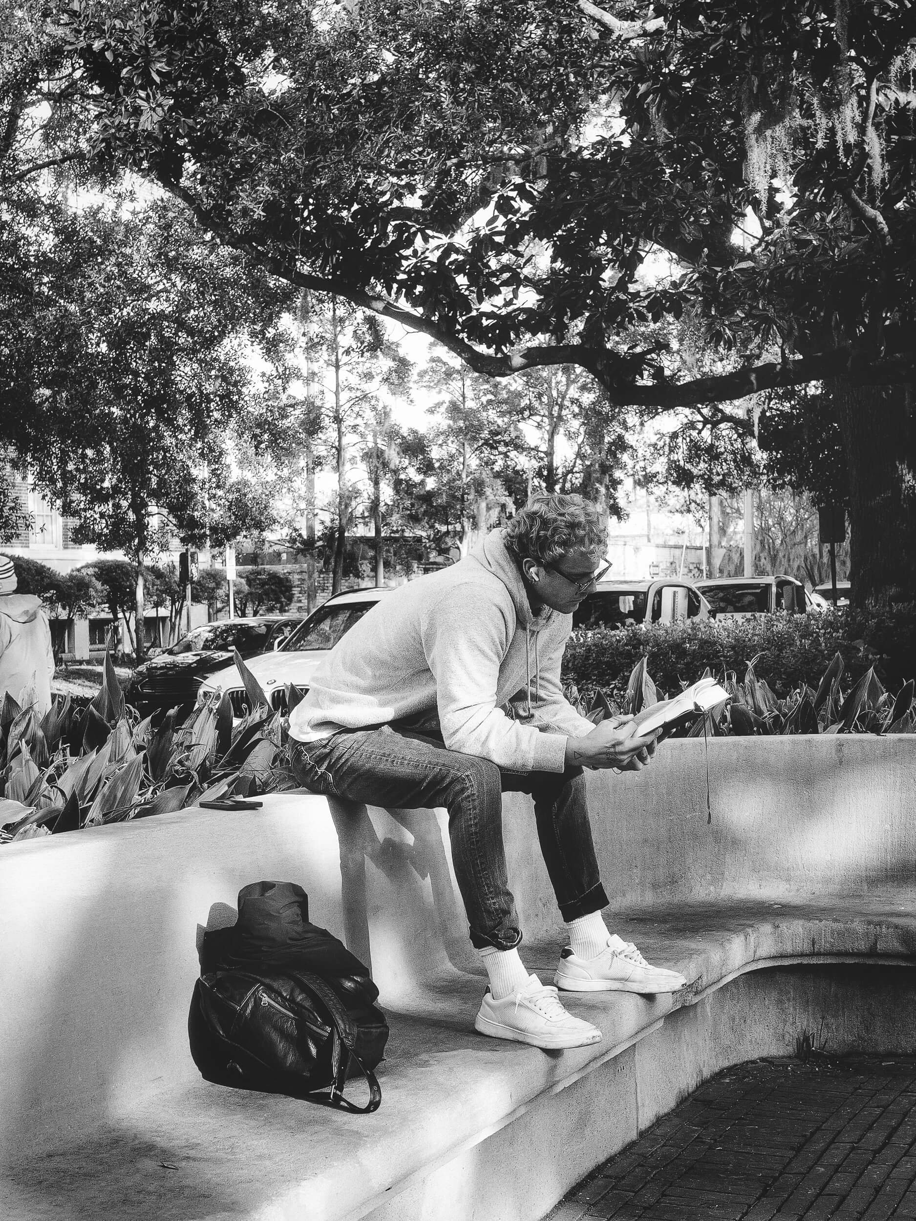 A solitary reader sits quietly on a curved stone bench beneath a sprawling oak draped in Spanish moss, immersed in a book amid the shaded calm of a Savannah square. The black-and-white tones highlight the textures of foliage, stone, and fabric, captu