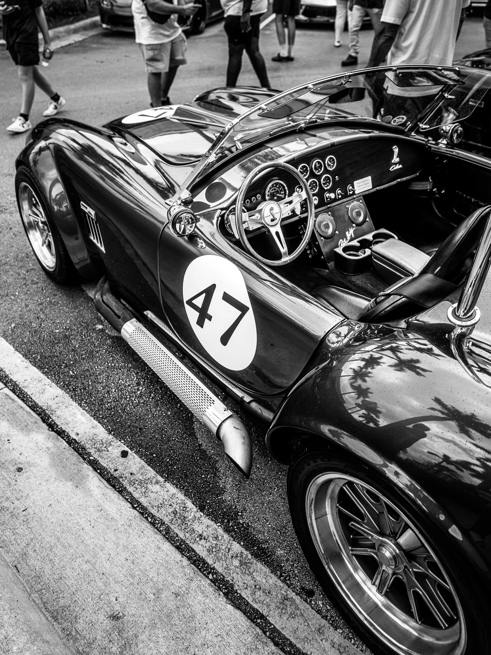 A classic Shelby Cobra roadster, bearing racing number 47, rests along the curb with its low, muscular stance and open cockpit on full display. In black and white, the image highlights the car’s iconic curves, side-mounted exhaust, and driver-focused