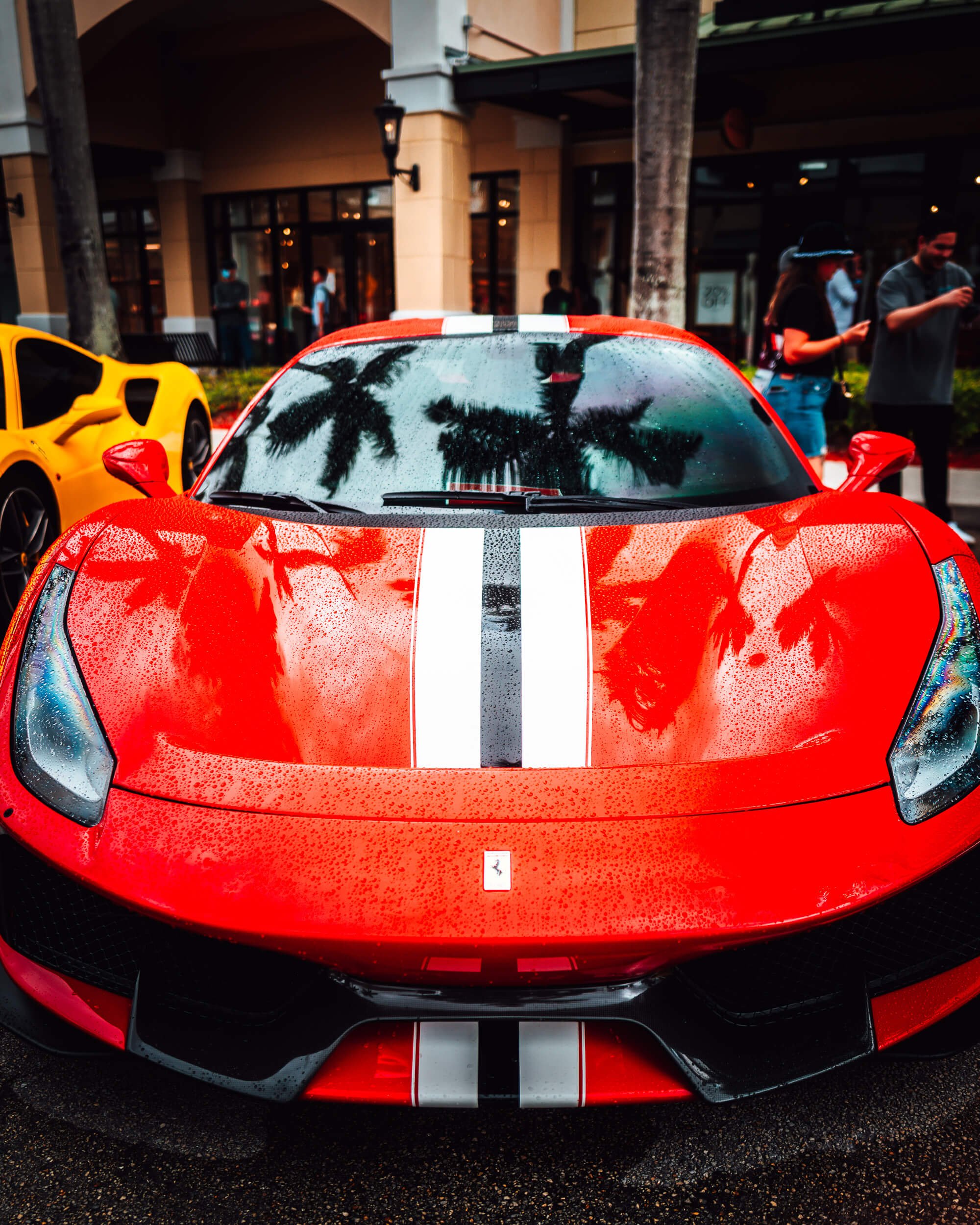 A vivid red Ferrari, streaked with twin racing stripes, commands attention as raindrops bead across its sculpted hood, reflecting swaying palm trees above. Parked amid a lively streetscape, the sleek supercar contrasts luxury and motion with the rela
