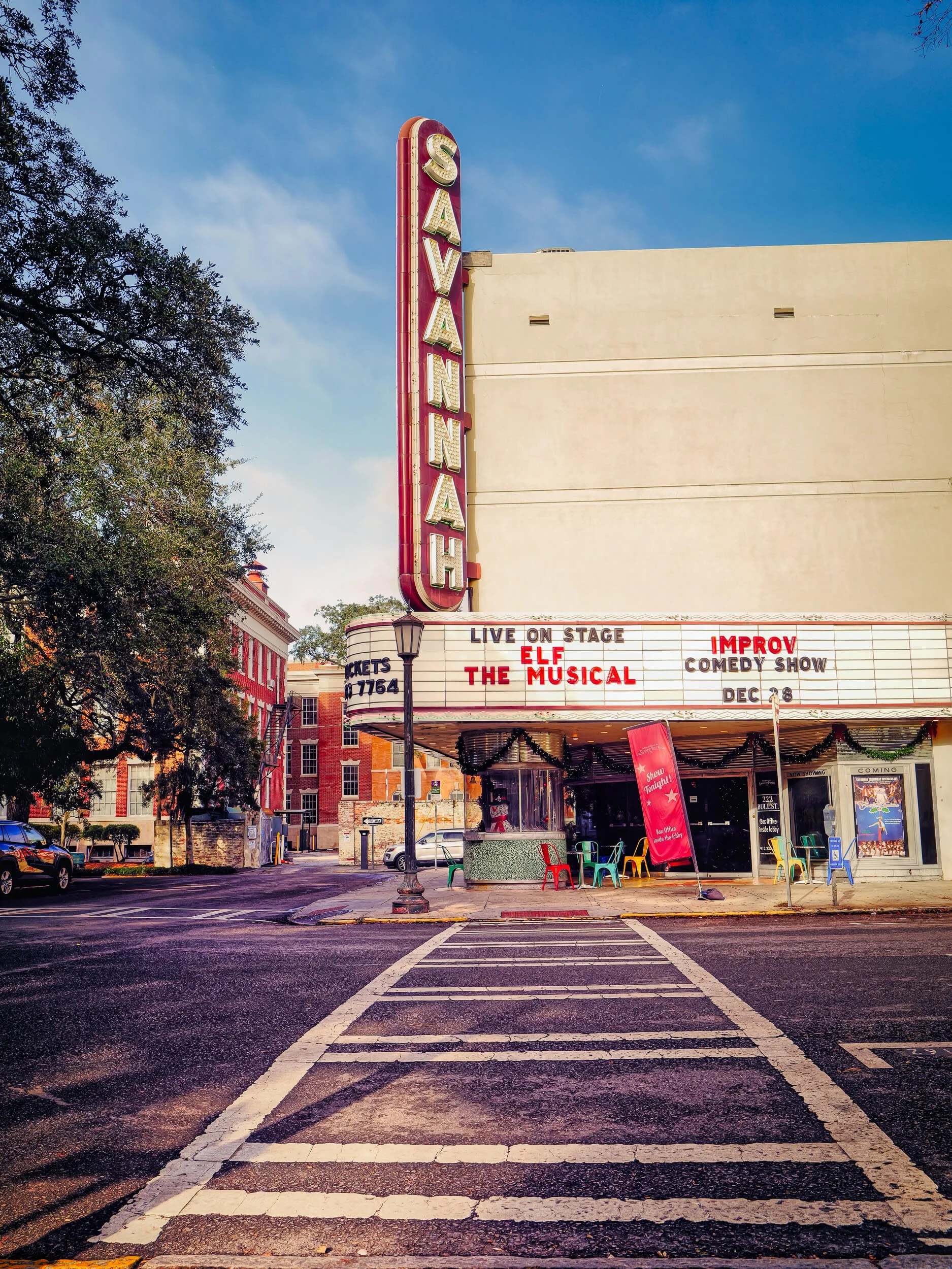 The historic Savannah Theatre stands prominently on a quiet street corner, its vintage marquee and vertical neon sign glowing with classic charm. Framed by a crosswalk in the foreground and bathed in warm daylight, the scene captures the timeless cha
